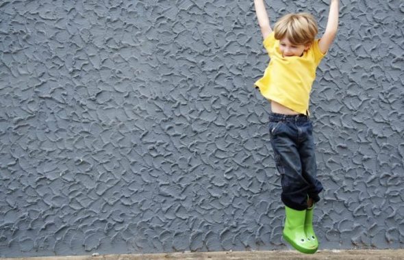 Pojke som hoppar upp i luften mot en grå bakgrund. Han har en illgul t-shirt på sig och gröna gummistövlar.