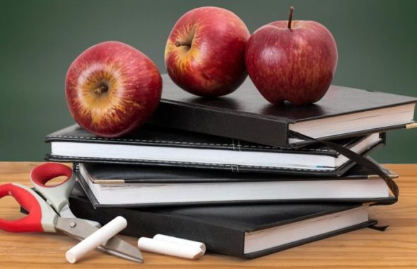 Böcker och äpplen i skolmiljö. Man ser också en sax och några kritor som är till en grön tavla i bakgrunden.